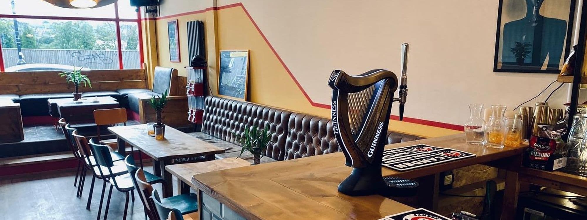 Cozy interior of Melodie 71 featuring a unique harp-shaped Guinness beer tap, wooden tables, tufted leather bench seating, and decorative plants, with warm lighting and framed artwork on the walls.