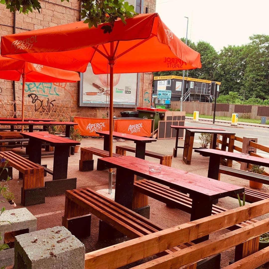 Outdoor seating area with wooden benches and tables under orange umbrellas at Melodie 71, with graffiti on a nearby wall and a street in the background.
