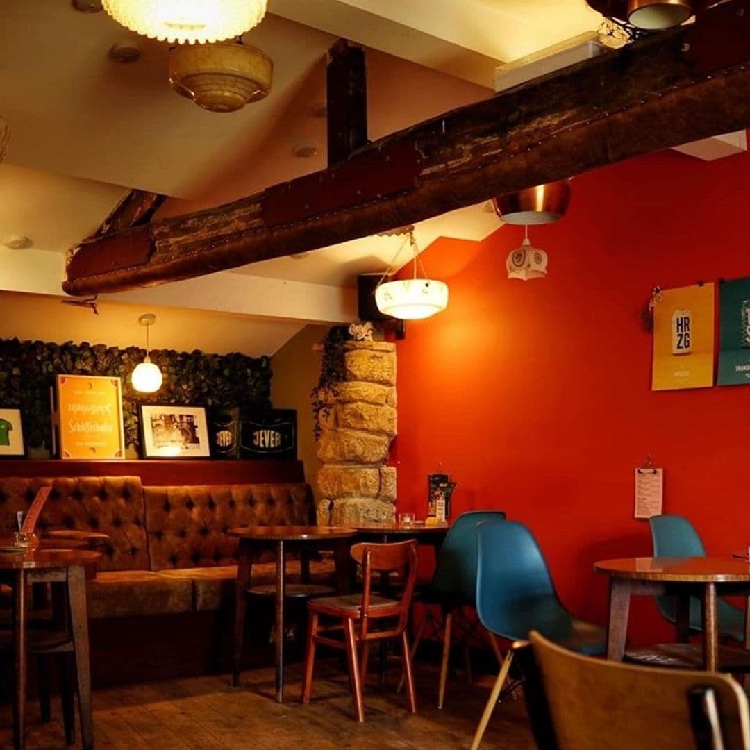 Cozy interior of North Bar Social with warm lighting, a mix of traditional wooden tables and chairs, a tufted leather sofa against a green hedge wall, and vibrant red walls adorned with framed artwork and posters.