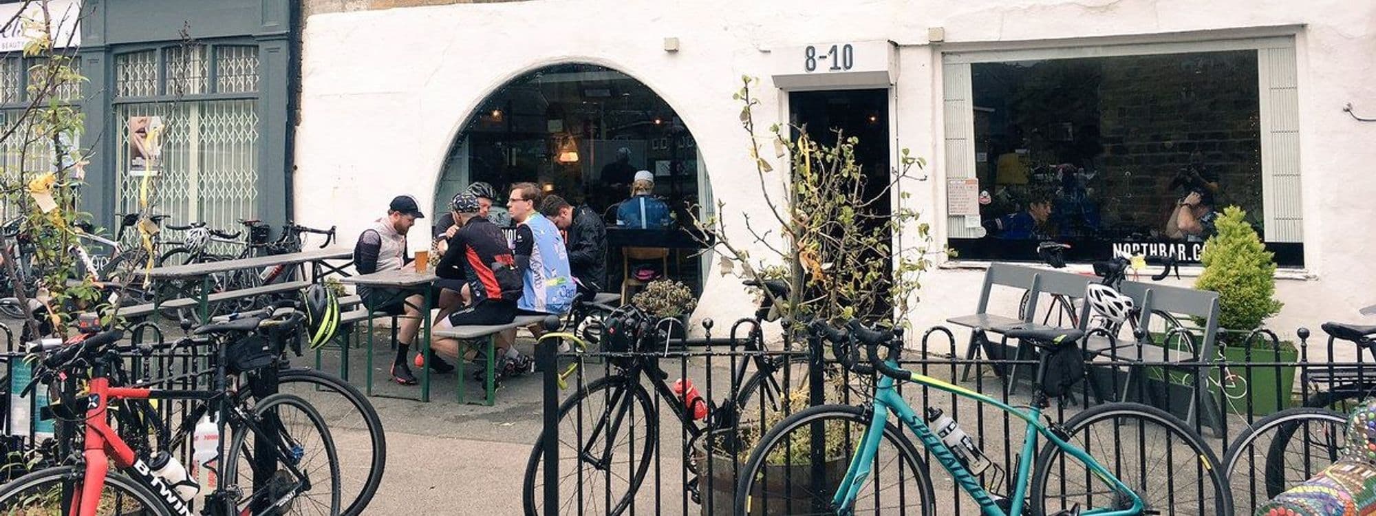 Outdoor seating area at North Bar Social with patrons enjoying drinks, surrounded by parked bicycles, in front of a white building with large windows and the establishment's name visible.