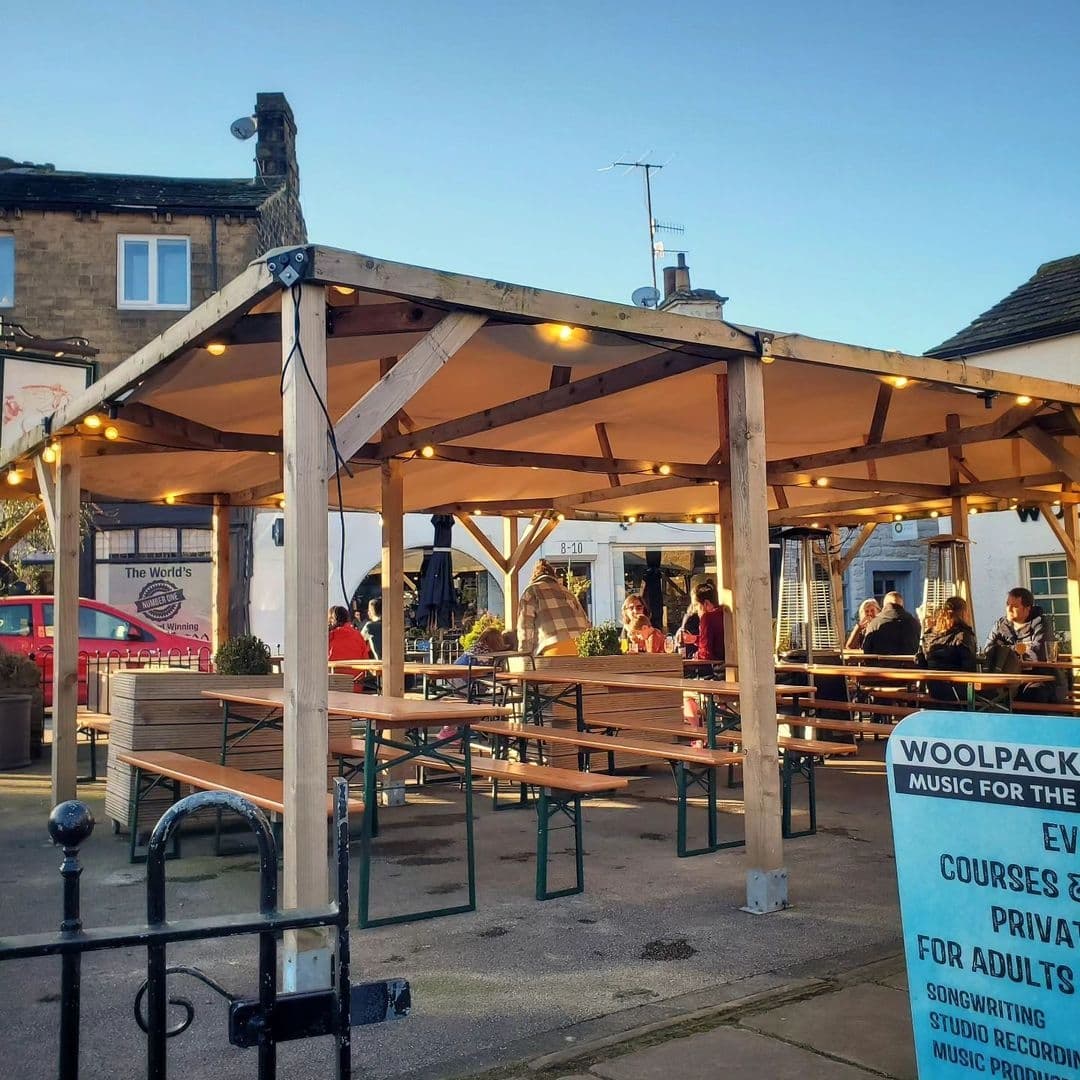 Outdoor seating area at North Bar Social with wooden tables and benches under a pergola with hanging lights, people socializing on a sunny day.