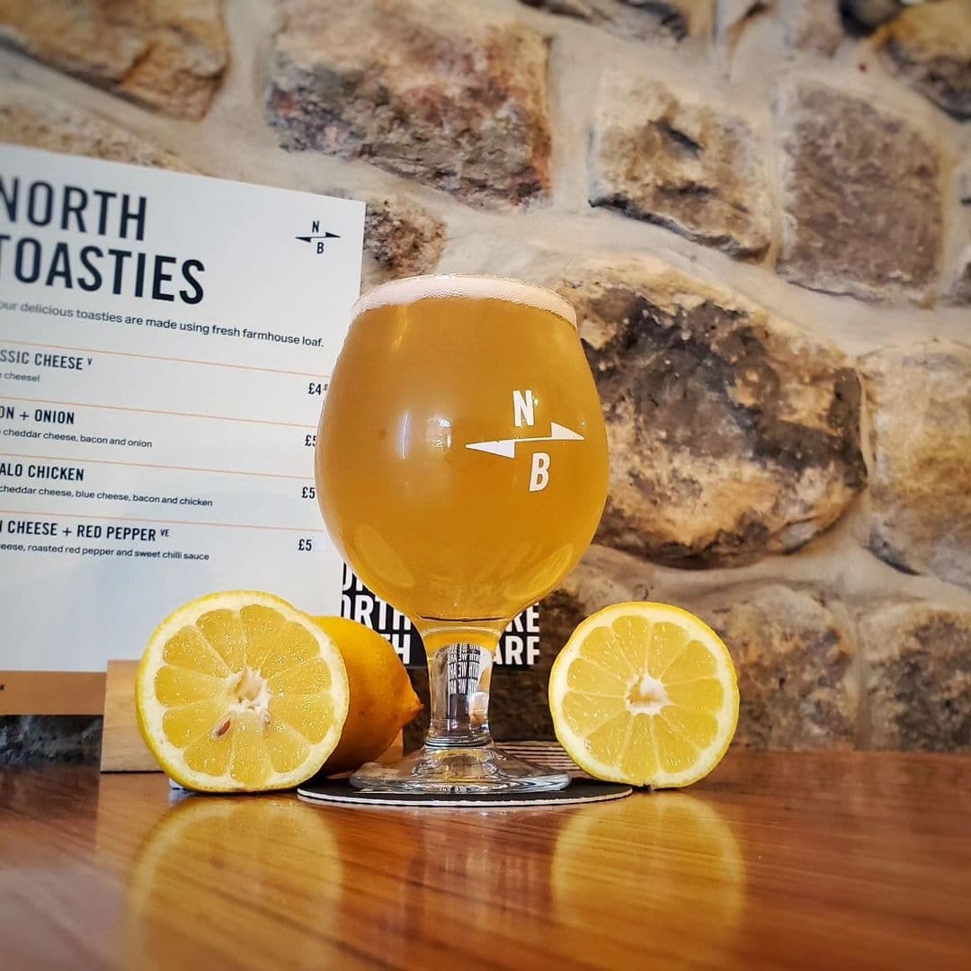 A refreshing glass of beer with the North Bar Social logo, accompanied by sliced lemons on a wooden bar top, with a menu in the background against a stone wall.