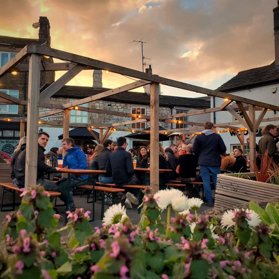 Outdoor seating area at North Bar Social with people socializing around wooden tables, string lights above, and flowers in the foreground at dusk.