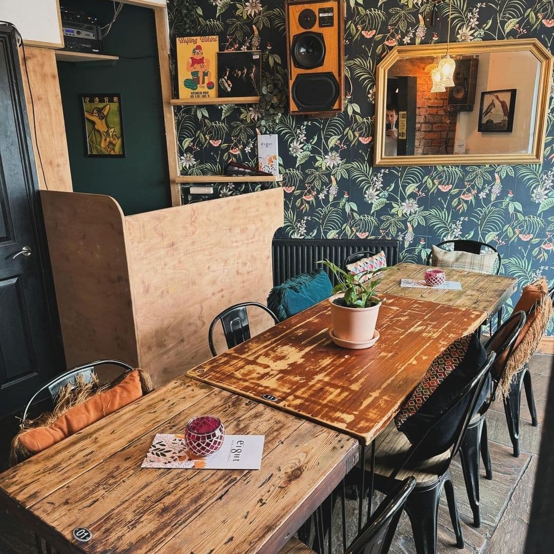 Interior of Number 8 featuring rustic wooden tables, eclectic decor with tropical wallpaper, framed art, and vintage speakers, creating a cozy and inviting atmosphere for visitors.