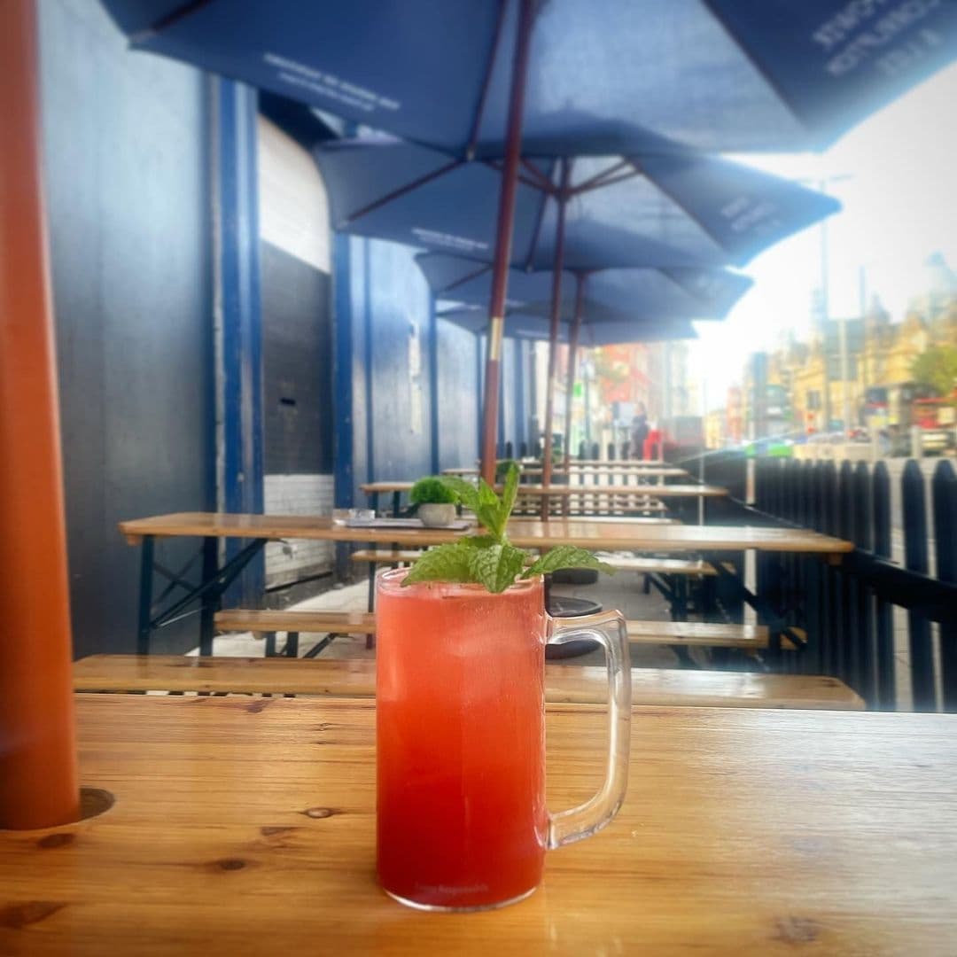 Refreshing red cocktail with mint garnish on wooden table at Power, Corruption & Lies, with outdoor seating and blue umbrellas in background.