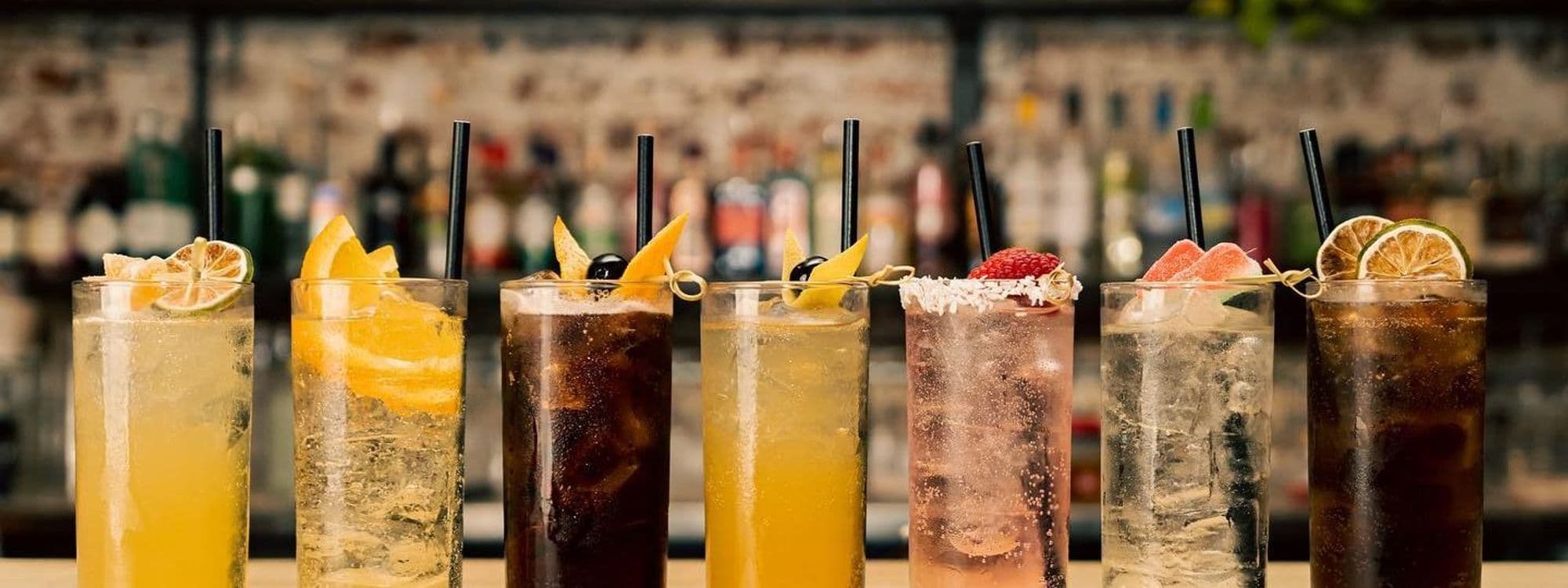 Assorted cocktails lined up on bar counter at Power, Corruption & Lies, with citrus garnishes and straws, inviting atmosphere for socializing.
