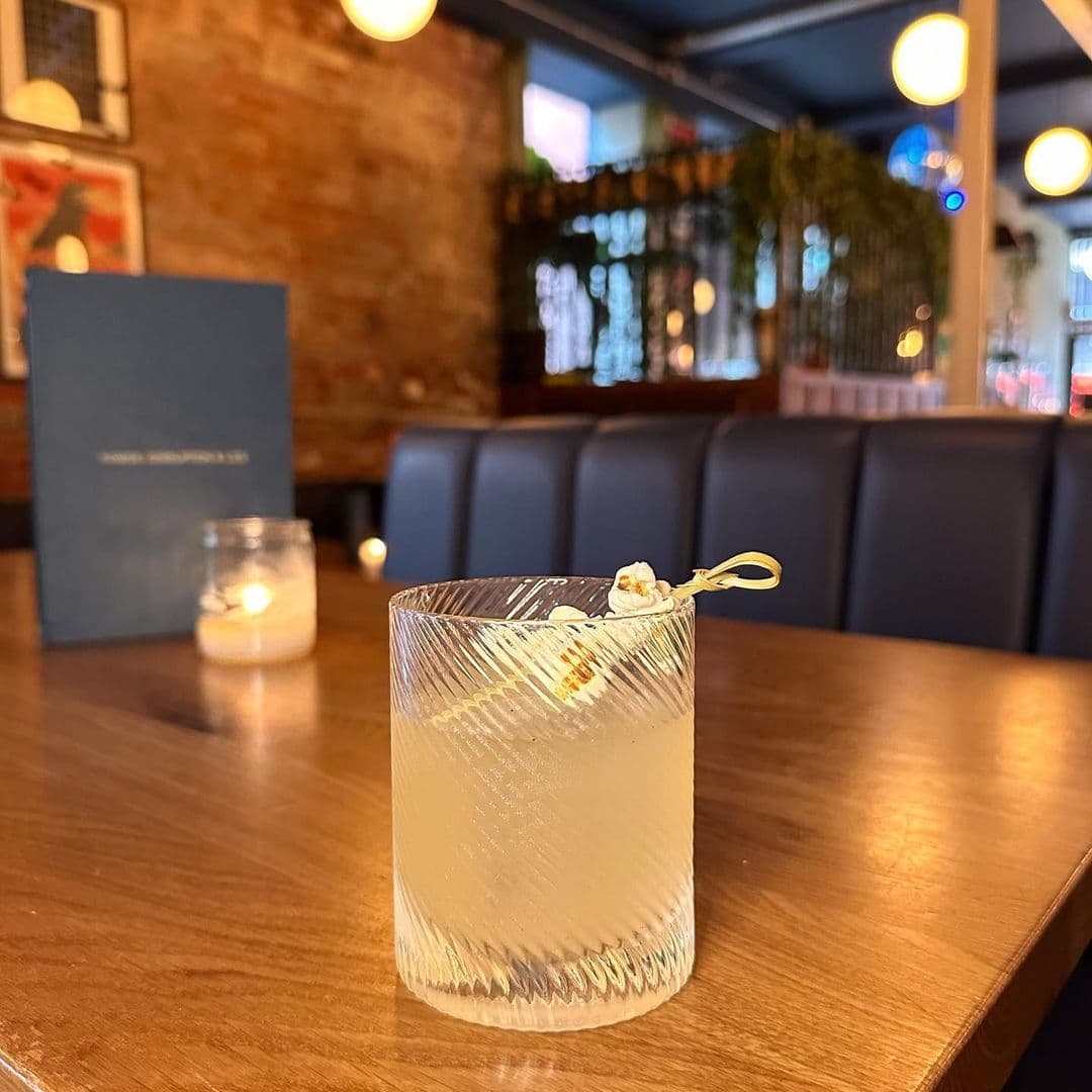 Craft cocktail in a textured glass garnished with popcorn at Power, Corruption & Lies, with cozy interior ambiance featuring warm lighting, exposed brick wall, and comfortable seating in the background.