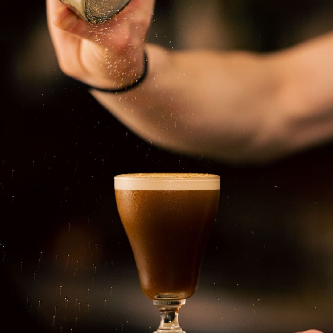 Hand sprinkling a dusting of spice over a frothy topped glass of dark beverage at Power, Corruption & Lies.