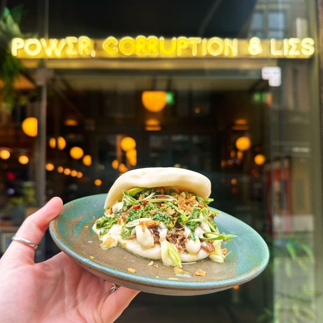 Hand holding a gourmet bao bun in front of the restaurant Power, Corruption & Lies with its glowing yellow neon sign.