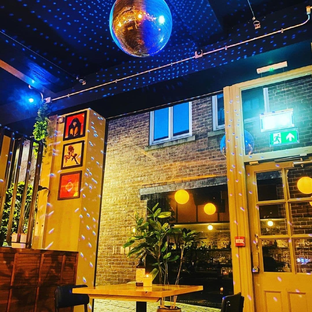 Alt text: "Cozy outdoor seating at Power, Corruption & Lies with warm lighting, a disco ball overhead casting dotted patterns, and vibrant framed artwork on the brick wall."