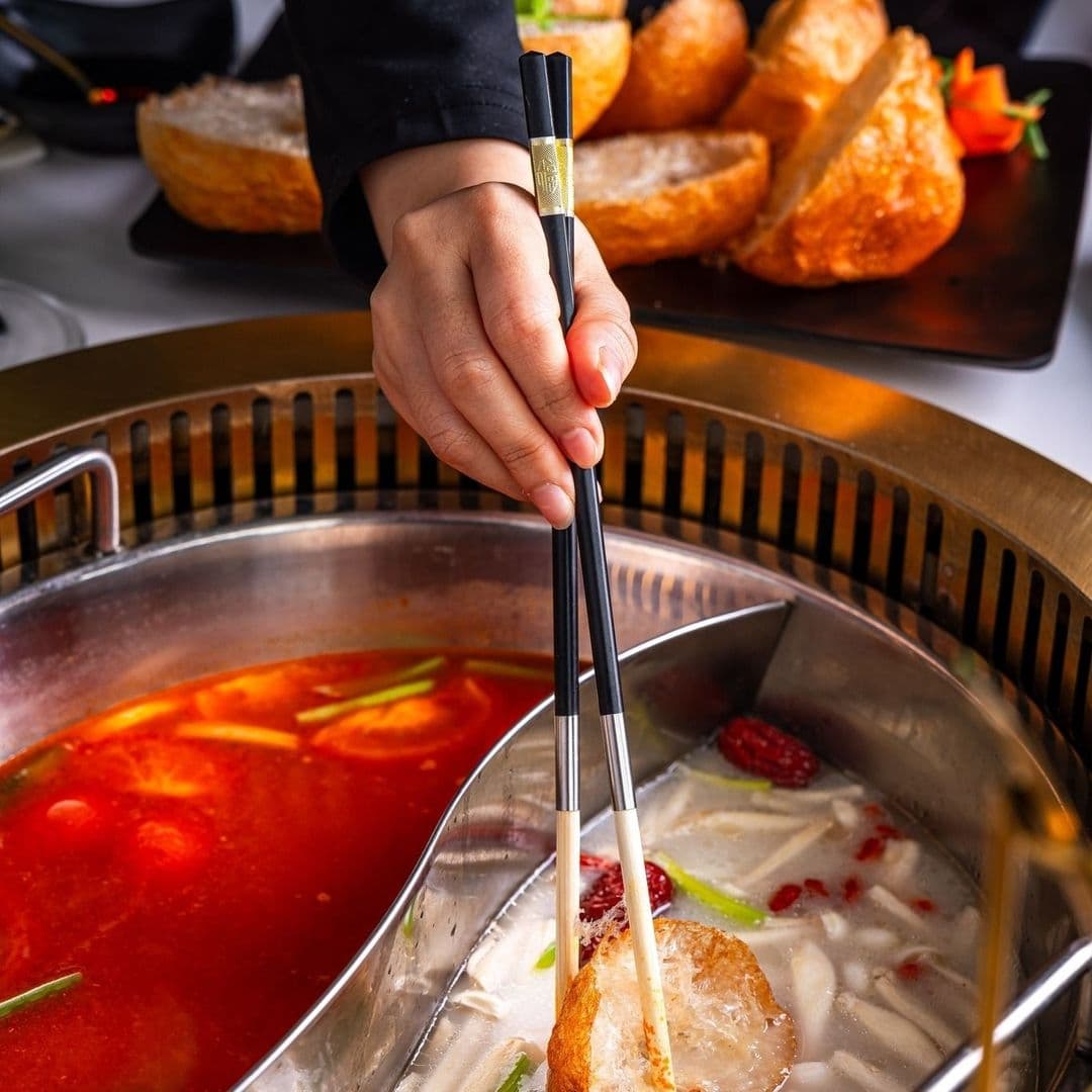 A person's hand using chopsticks to dip a piece of food into a split hot pot with both spicy and clear broth at TAO.