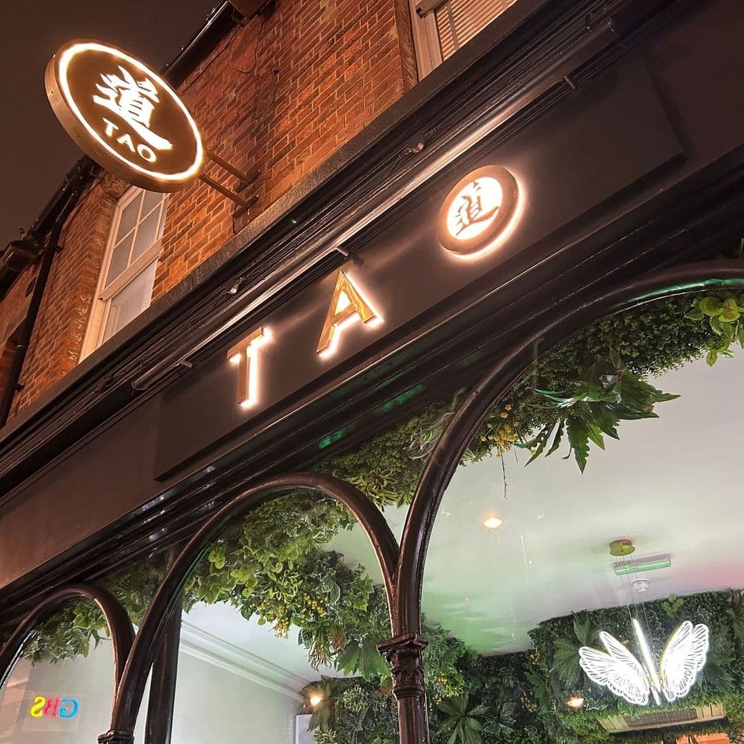 Exterior view of TAO with illuminated signage above a window framed by green foliage and a neon butterfly decoration visible through the glass.