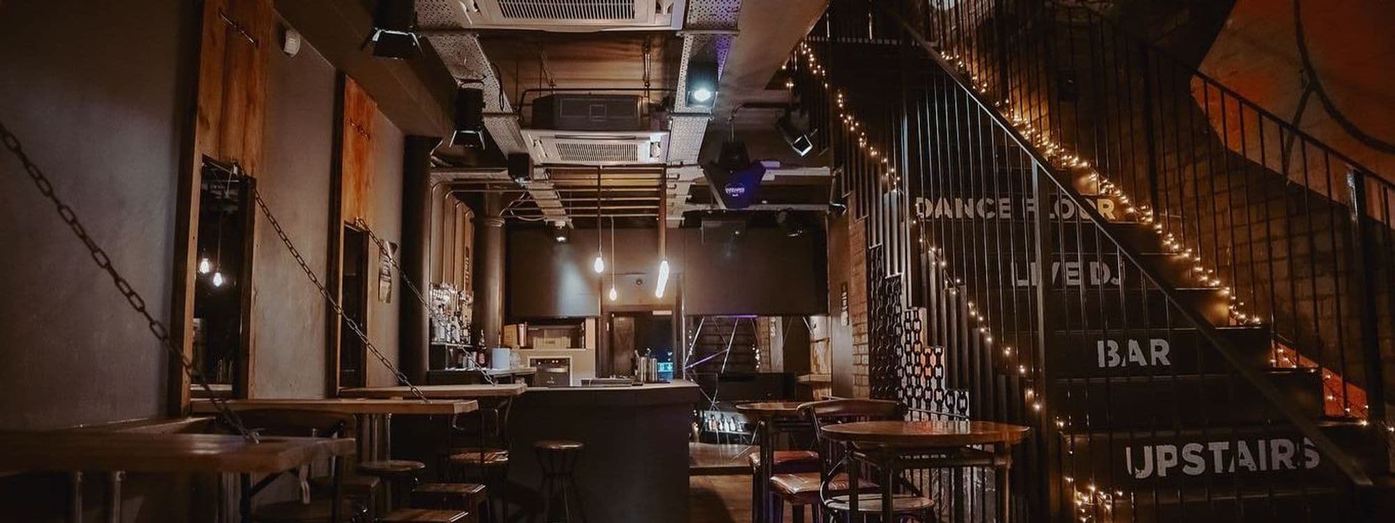 Interior of Ten Bar with dim lighting, featuring wooden tables, a bar counter, hanging industrial lights, and a staircase leading upstairs adorned with string lights.