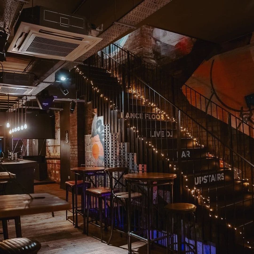 Interior of Ten Bar with ambient lighting, featuring a staircase with fairy lights, high stools at the bar, and a dance floor sign.