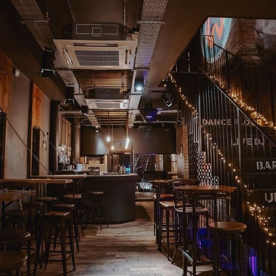 Interior of Ten Bar with dim lighting, featuring a central bar area surrounded by wooden stools, a staircase with illuminated 'DANCE FLOOR' sign, and exposed brick walls.