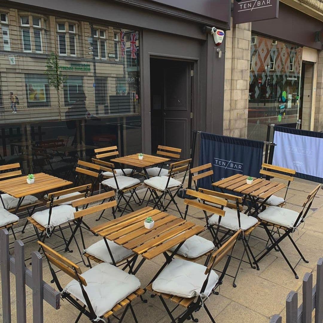Outdoor seating area at Ten Bar with wooden tables and chairs with white cushions, ready for guests.