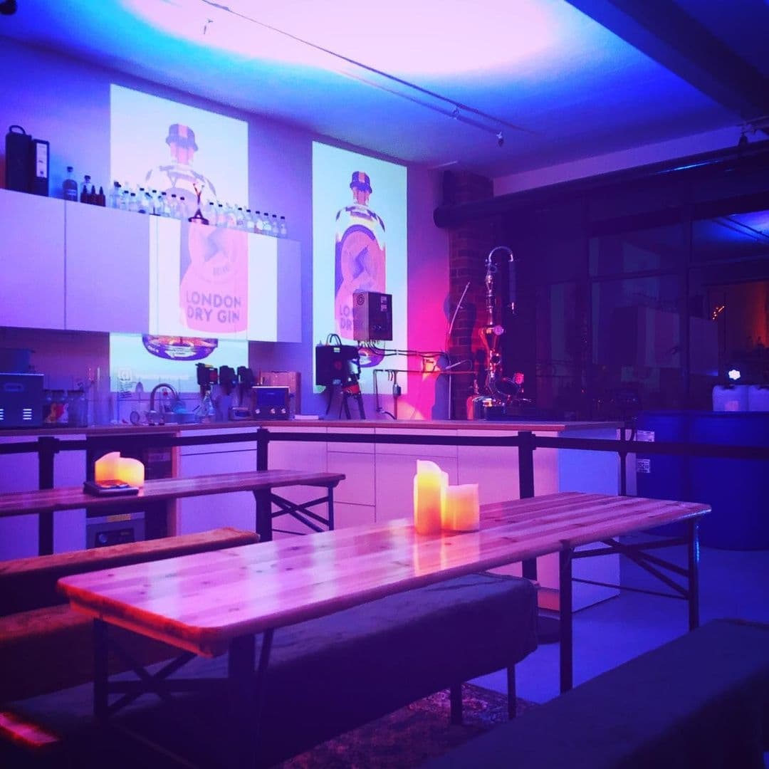 Interior of Test Bar with ambient blue lighting, wooden tables, and benches. Projected images of London Dry Gin on the wall above the bar counter, with lit candles on tables creating a cozy atmosphere.