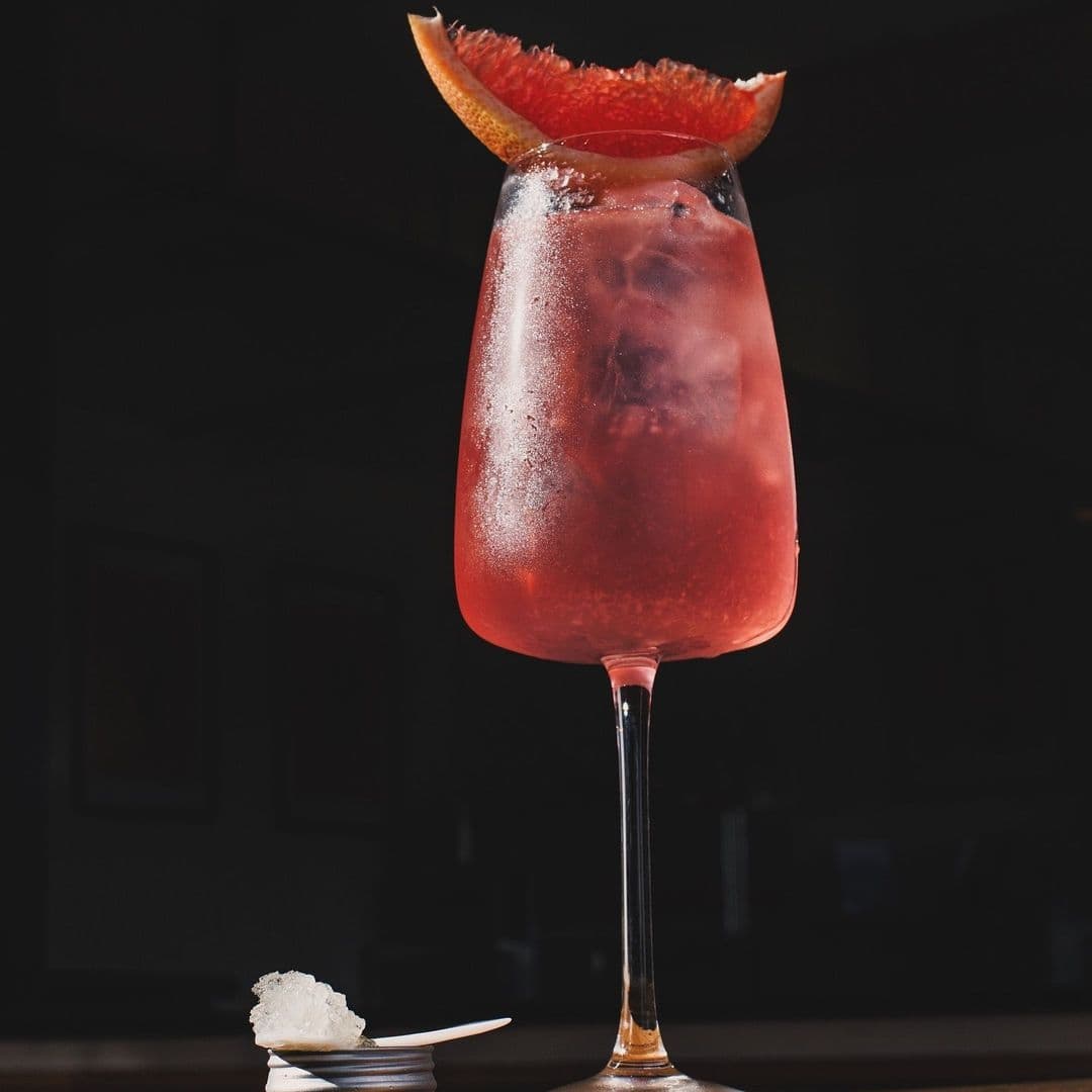 Refreshing cocktail served at Test Bar, featuring a tall glass with a vibrant red beverage, ice cubes, and a slice of orange garnish on the rim, presented on a dark backdrop with a small dish of appetizer.