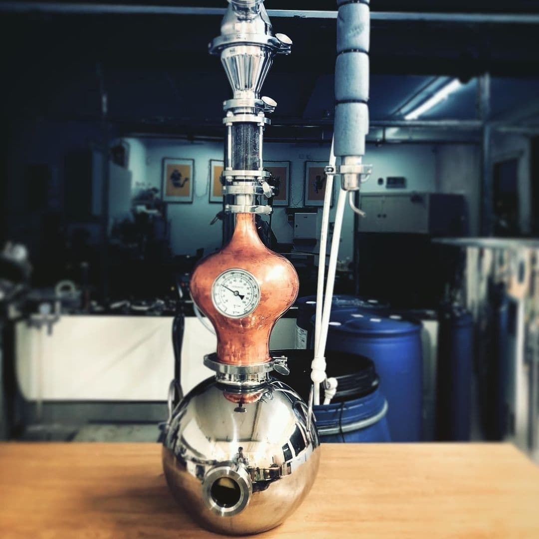 Copper and stainless steel distillation apparatus at Test Bar, with a pressure gauge and various connecting tubes, set against a blurred background of an industrial setting.