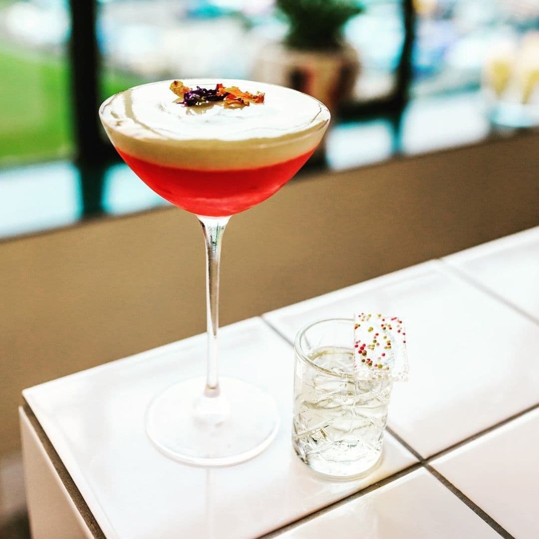Elegant cocktail with creamy top and red base served in a stemmed glass, accompanied by a clear drink with ice in a tumbler, on a white table at Test Bar.