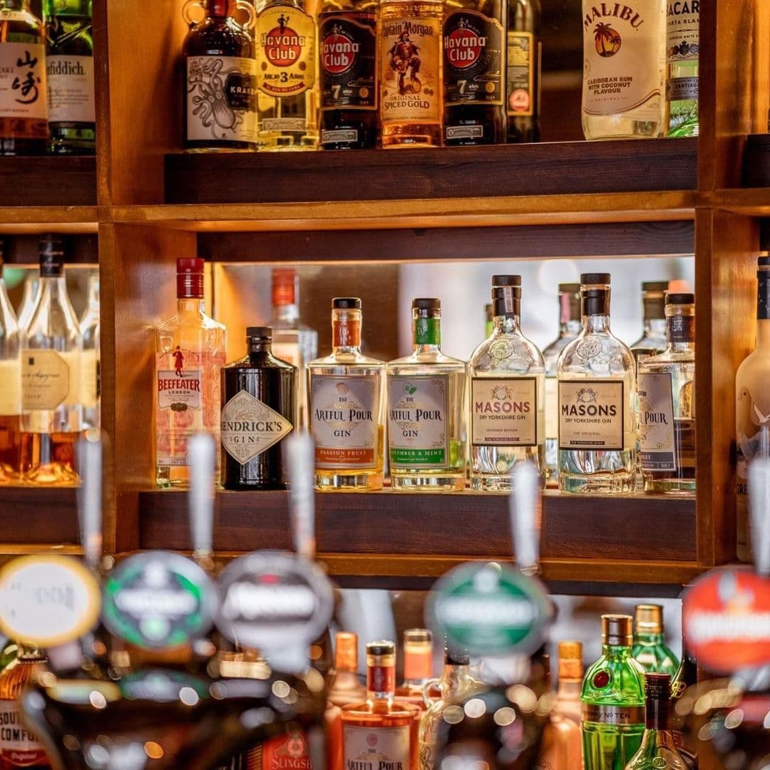 A variety of spirits and liquor bottles on wooden shelves at Tharavadu bar, with focus on gin and rum brands, and blurred beer taps in the foreground.