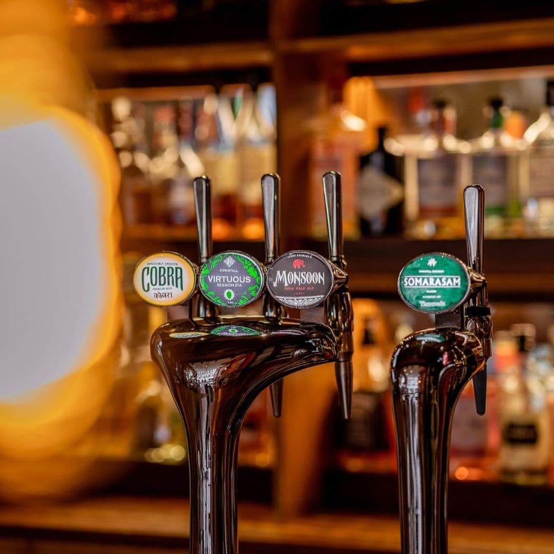 Beer taps with various labels including Cobra and Monsoon at Tharavadu bar, with blurred background of bottles on shelves.