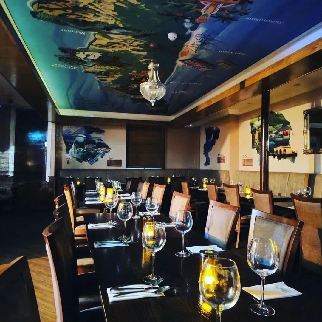 Elegant dining setup at Tharavadu with tables arranged with glassware and cutlery, under a ceiling adorned with a large, artistic map mural.