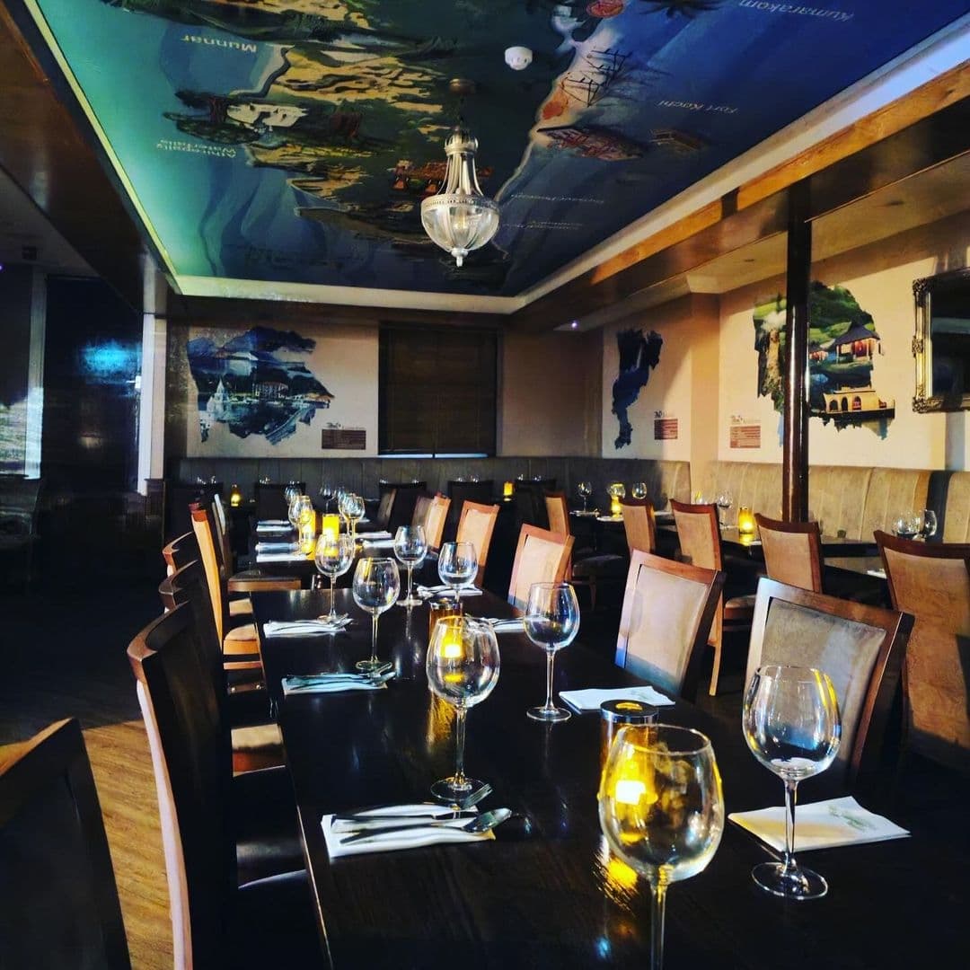 Elegant dining setup at Tharavadu with tables adorned with wine glasses and cutlery under warm ambient lighting and a ceiling painted with cultural artwork.