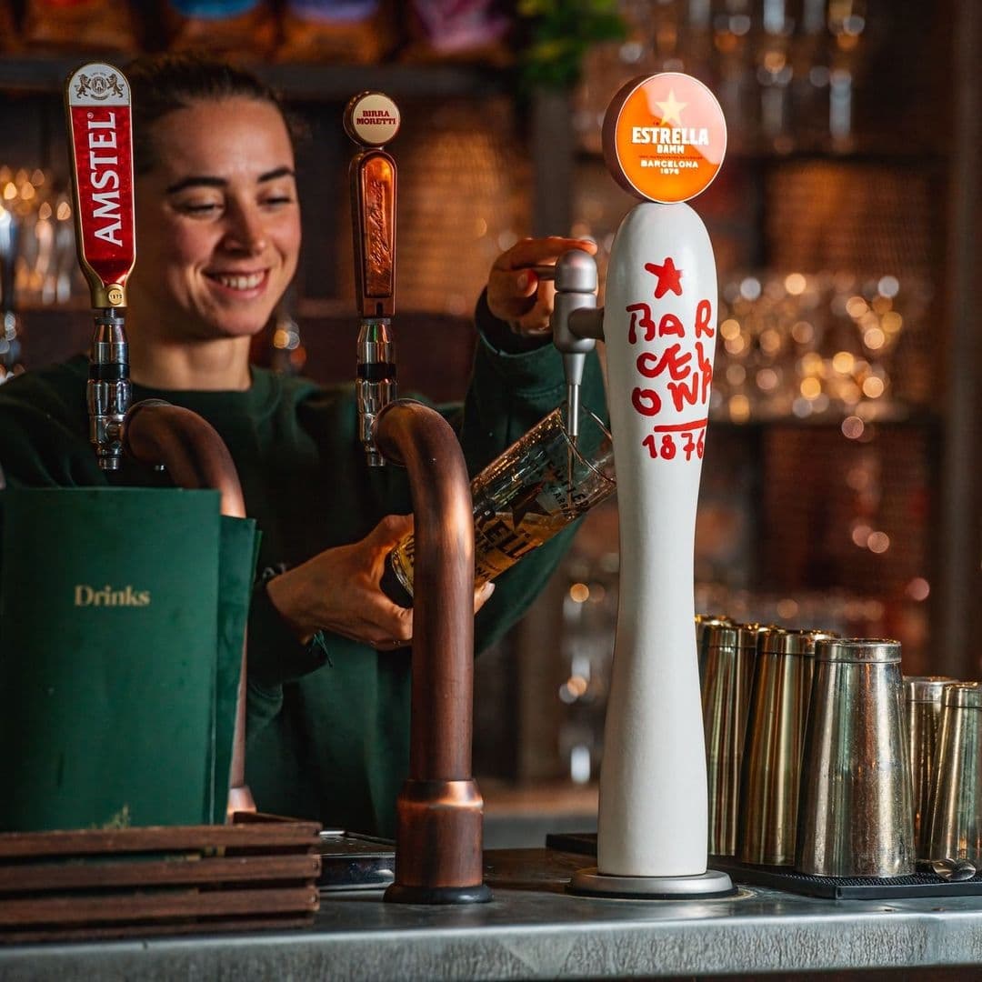 A smiling bartender pouring a drink at The Botanist Trinity with beer taps featuring Amstel and Estrella in the foreground.