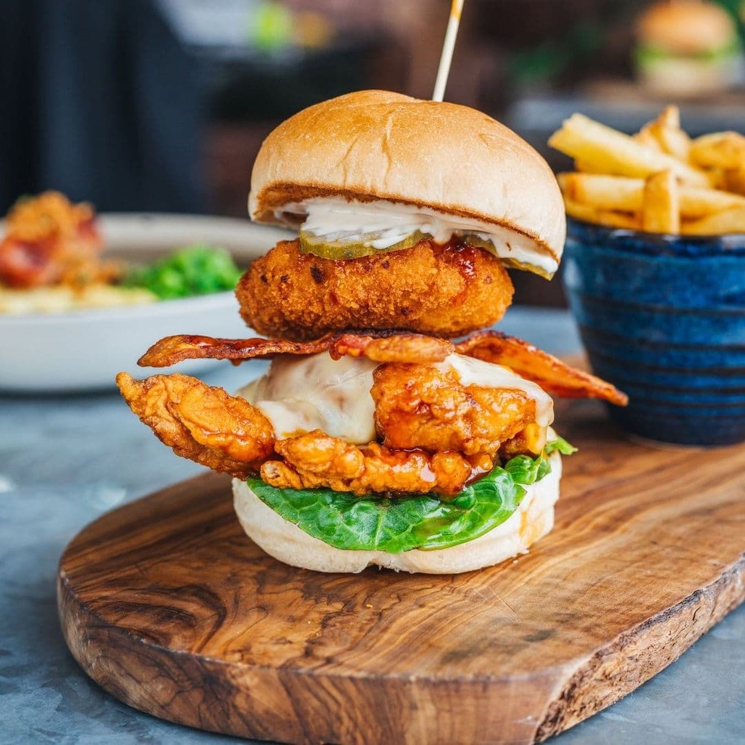 Gourmet chicken burger with crispy bacon, cheese, and spicy sauce served on a wooden board at The Botanist Trinity, with a side of fries in a blue bowl.