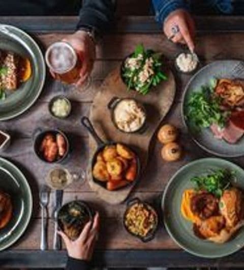 Diverse selection of gourmet dishes and drinks on a wooden table at The Botanist Trinity, with people dining and sharing a meal.