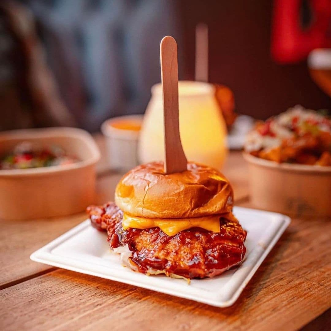 Cheeseburger with a wooden skewer on a square plate at The Brewery Tap, with blurred background of additional dishes and a warm ambiance.