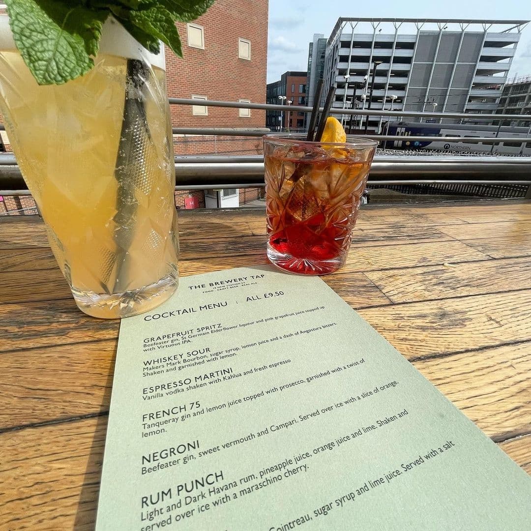 Cocktails on a wooden table at The Brewery Tap with a cocktail menu in view, featuring a tall glass of Grapefruit Spritz and a short glass of Negroni, against an urban backdrop with buildings and a clear sky.