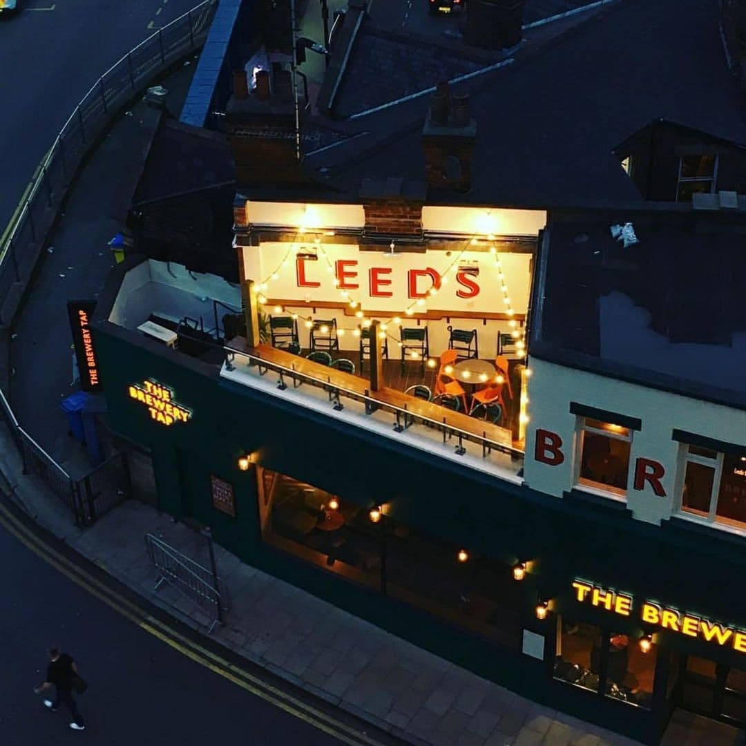 Aerial view of The Brewery Tap at dusk, featuring outdoor seating with string lights and a large 'LEEDS' sign illuminated above the entrance.
