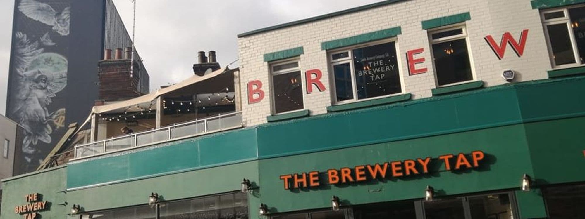 Exterior view of The Brewery Tap, featuring a white brick building with large red letters spelling "BREW" on the upper level, and the establishment's name in orange letters over the entrance. A covered outdoor seating area is visible on the upper deck, adorned with string lights.