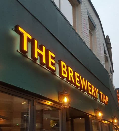Exterior view of The Brewery Tap with illuminated signage and warm lighting from hanging lamps at dusk.