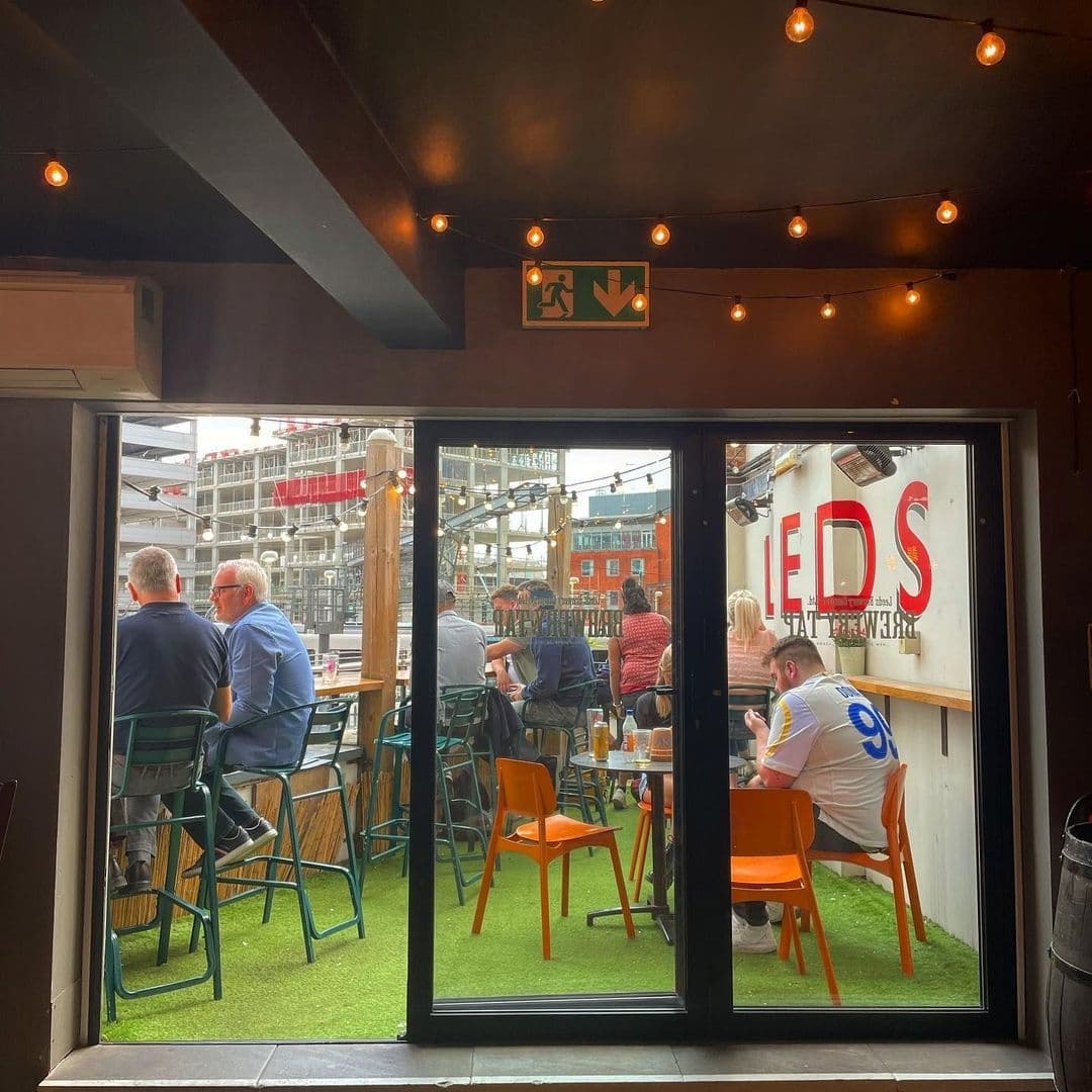 View from inside The Brewery Tap looking out to a lively outdoor seating area with patrons enjoying drinks, surrounded by urban construction and string lights above.