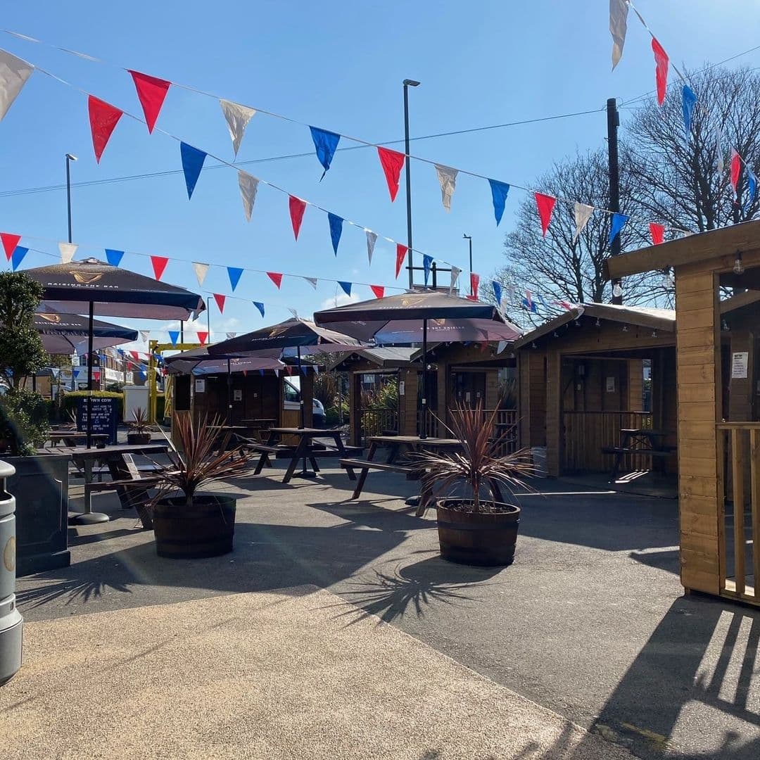 Outdoor seating area at The Brown Cow with wooden tables, umbrellas, and colorful bunting under a clear blue sky.