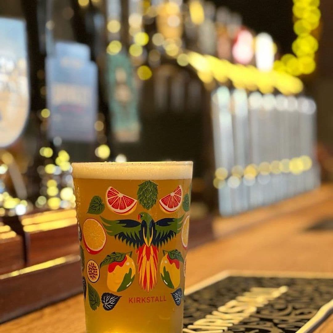 A pint of beer with a colorful label featuring a bird and citrus fruits, served at The Cross Keys pub, with a blurred background of the bar interior.