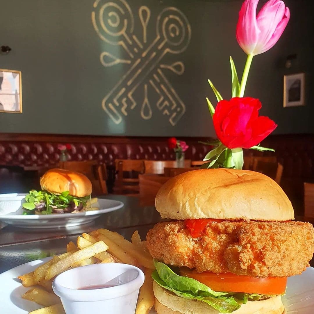 Delicious crispy chicken burger with fries served at The Cross Keys, with a vibrant red tulip in the foreground adding a touch of elegance to the dining experience.