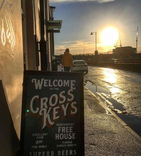 Sunset view outside The Cross Keys with a prominent signboard featuring the pub's name, advertising excellent food, fine wines, and superb beers, with a person walking away in the background.