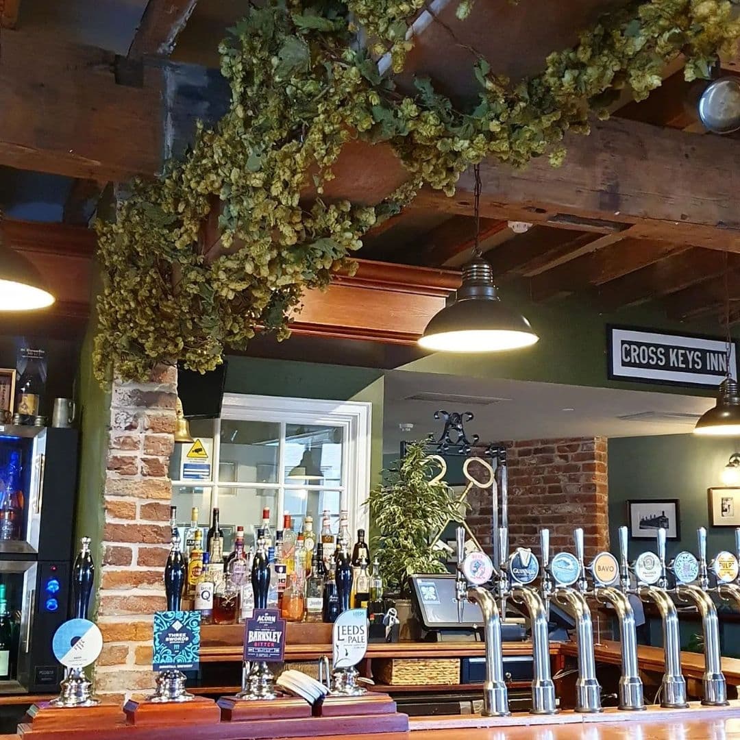Interior view of The Cross Keys showing a traditional bar with a variety of beer taps, exposed brick walls, and hops hanging from wooden beams.