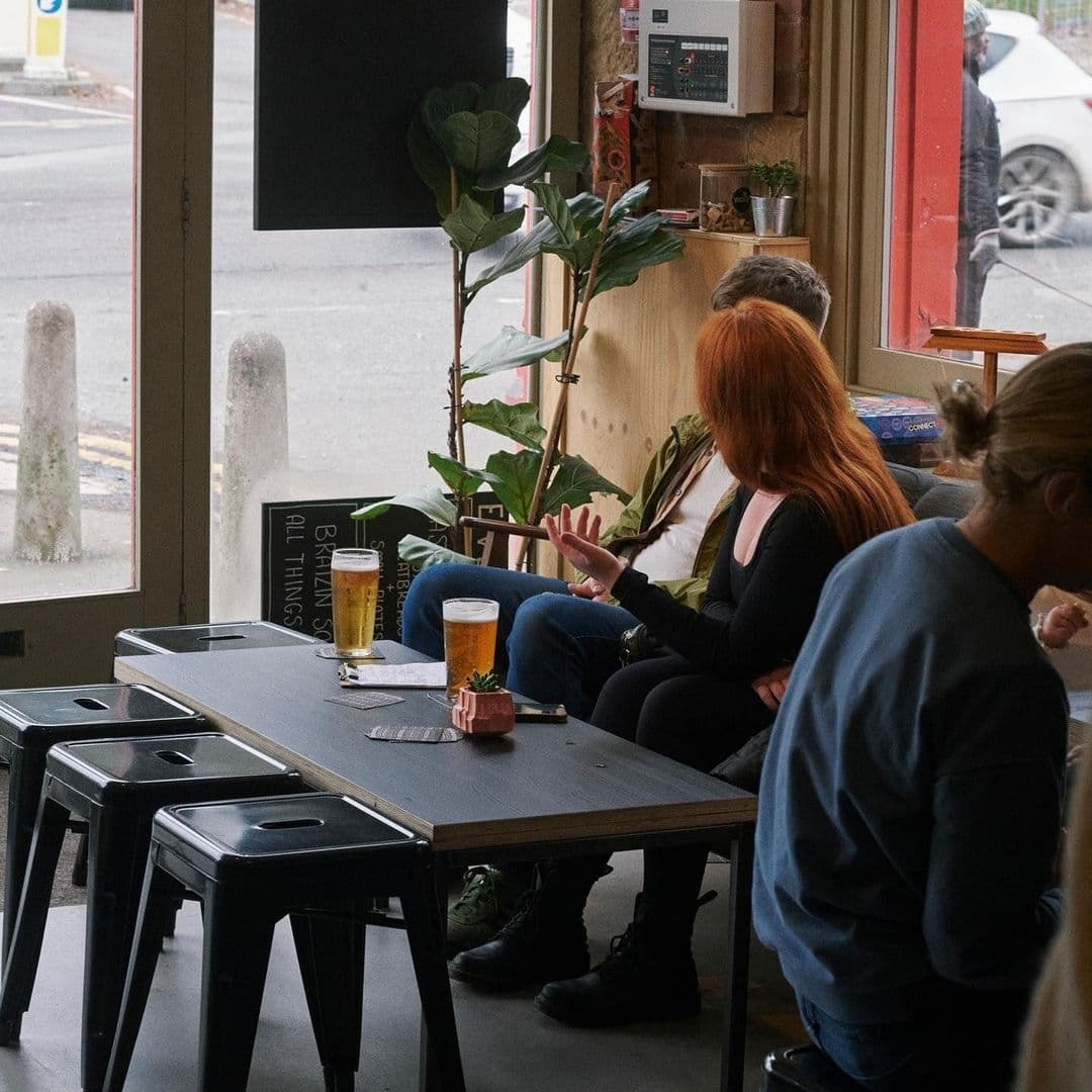 Casual indoor setting at The Melbourne with patrons enjoying drinks and conversation at a wooden table near an open window, with a view of the street outside.