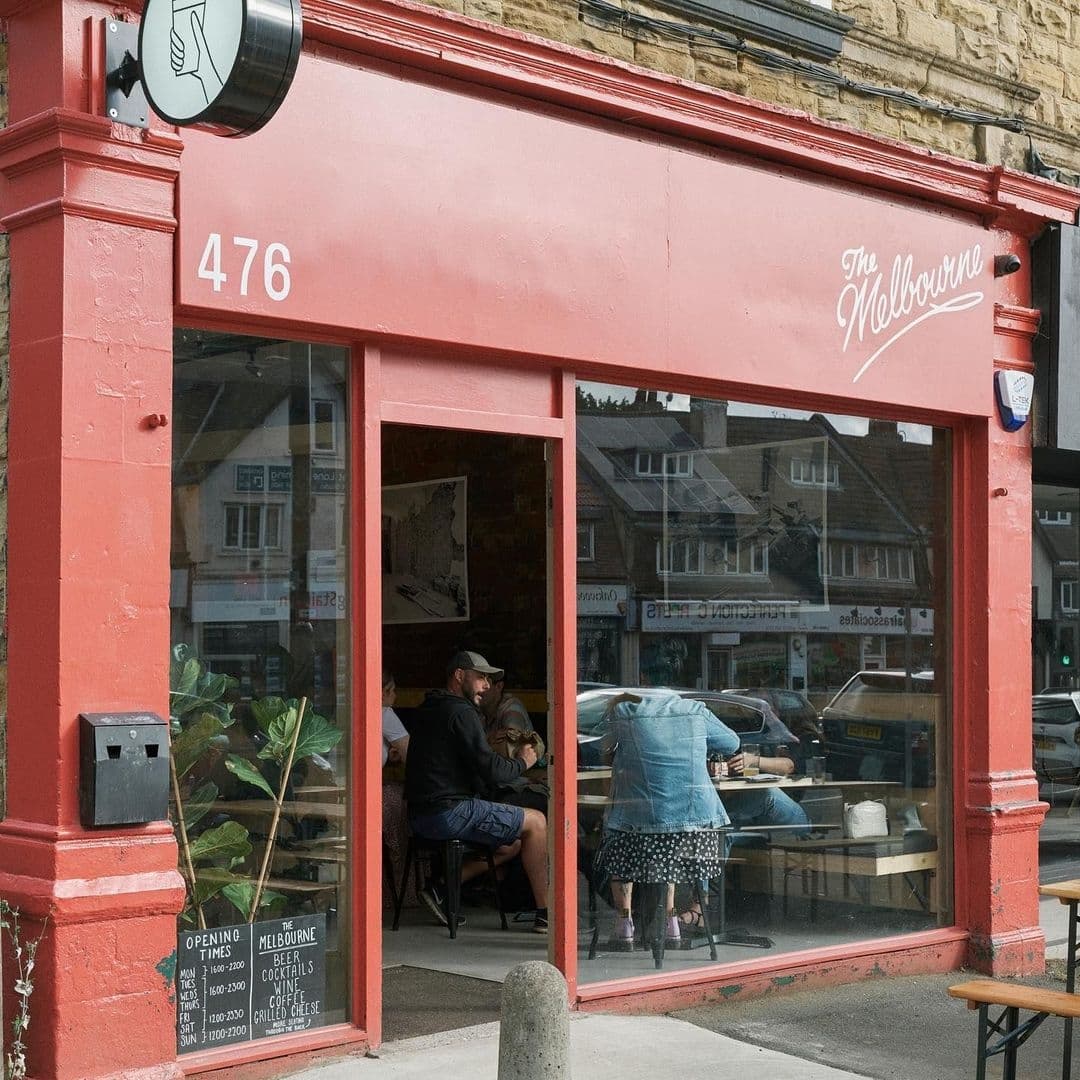 Exterior view of The Melbourne cafe with patrons dining inside, red storefront on a sunny day with street reflection in the windows.