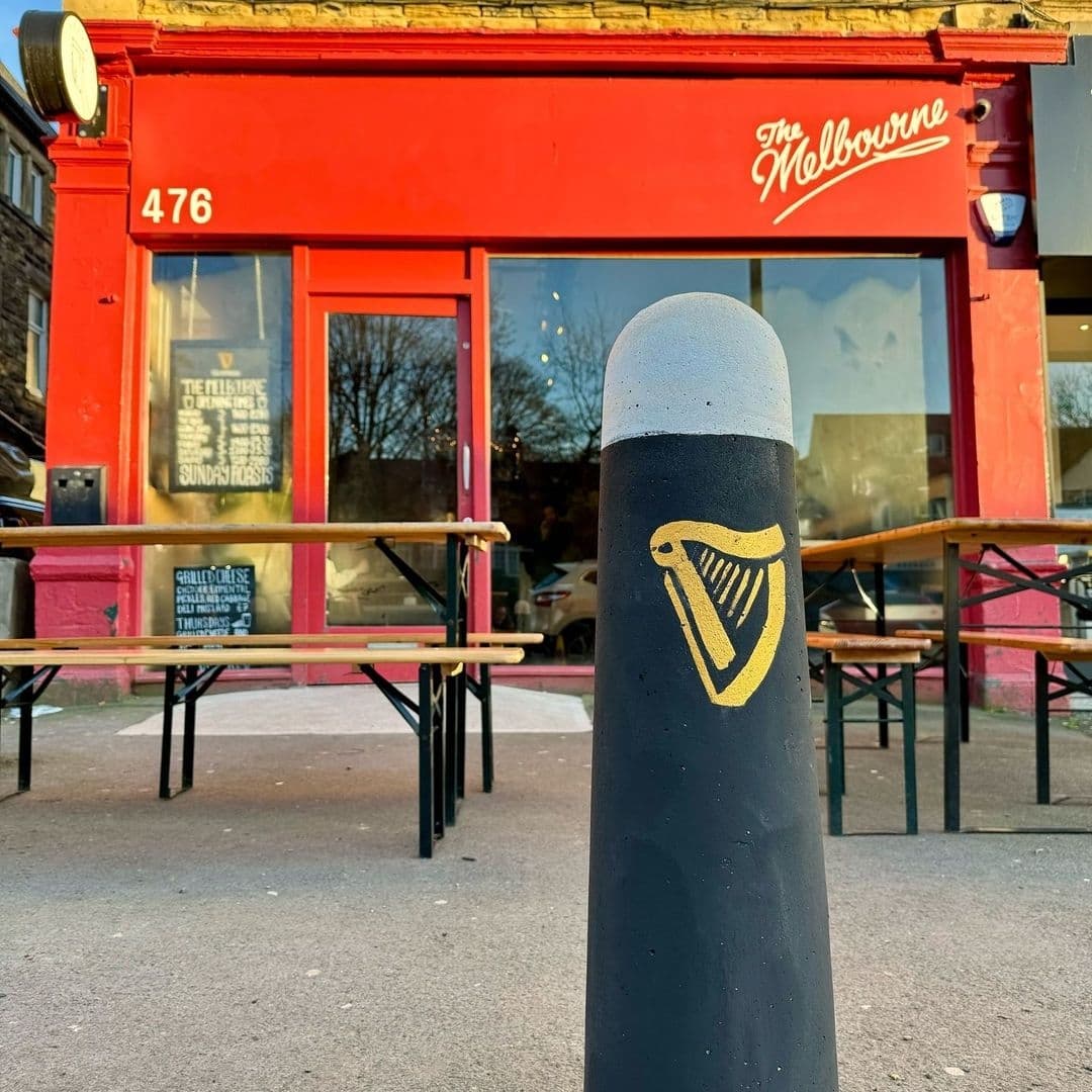Exterior view of The Melbourne with vibrant red facade, signage above the entrance, and outdoor seating area with wooden benches. A black bollard with a harp symbol in the foreground.