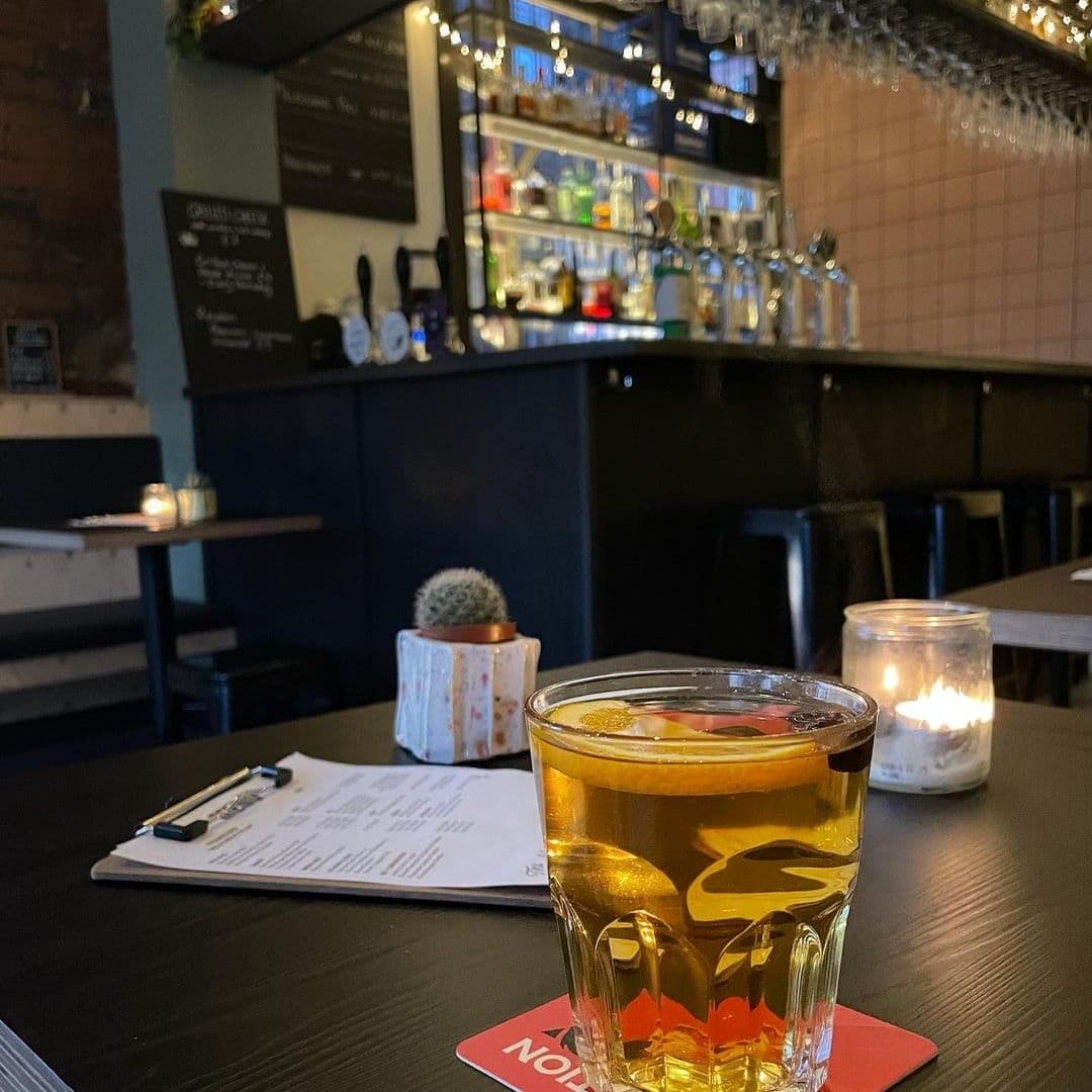 Cozy interior of The Melbourne featuring a close-up of a pint of beer on a table with a menu, lit candle, and a small cactus, with a blurred background of a bar shelf with various bottles and glasses.