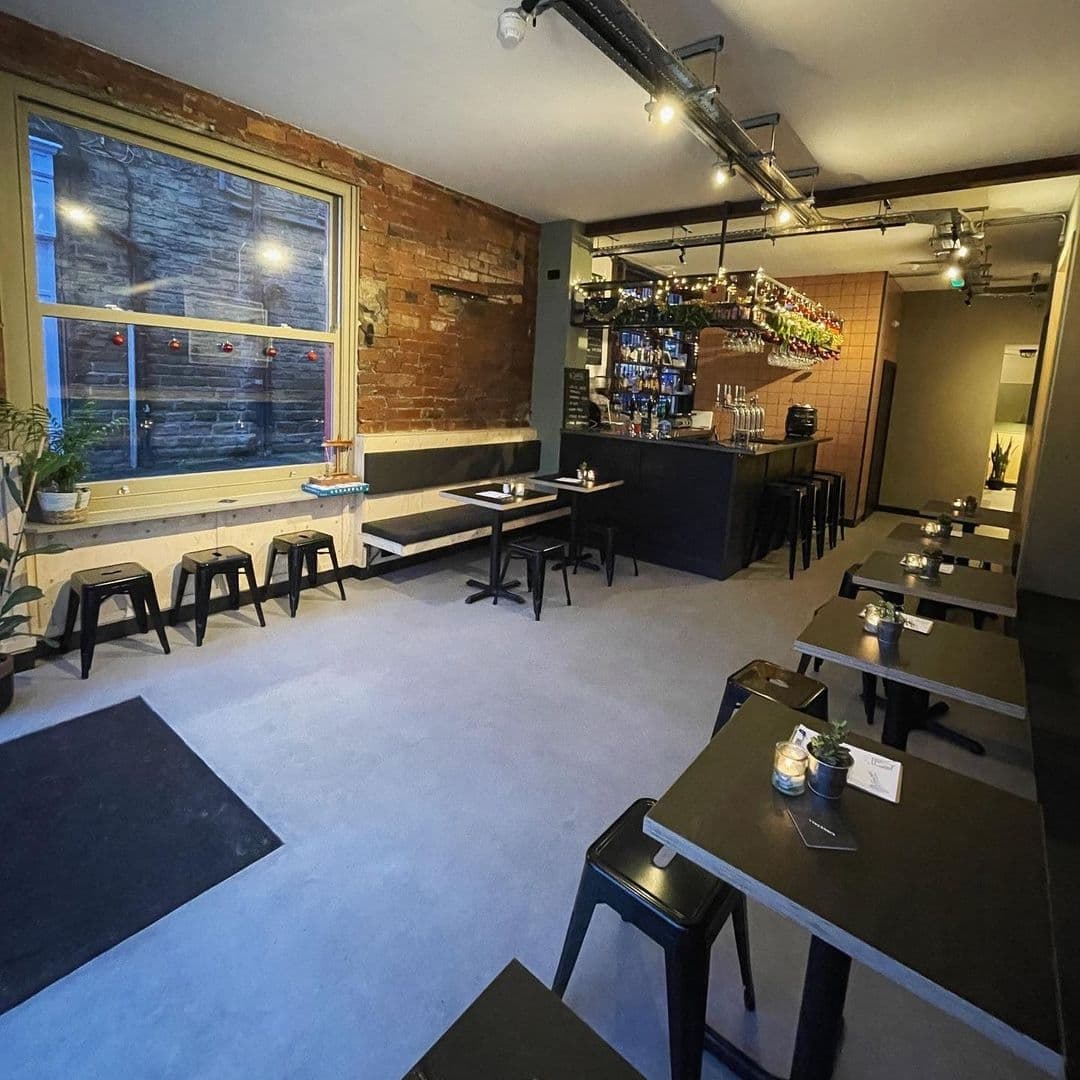 Interior of The Melbourne featuring a cozy dining area with exposed brick walls, simple black furniture, and a well-lit bar with hanging wine glasses.