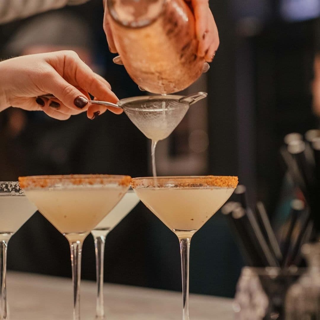 Bartender pouring a cocktail through a strainer into a martini glass with two more glasses in the foreground, at The Turk’s Head.