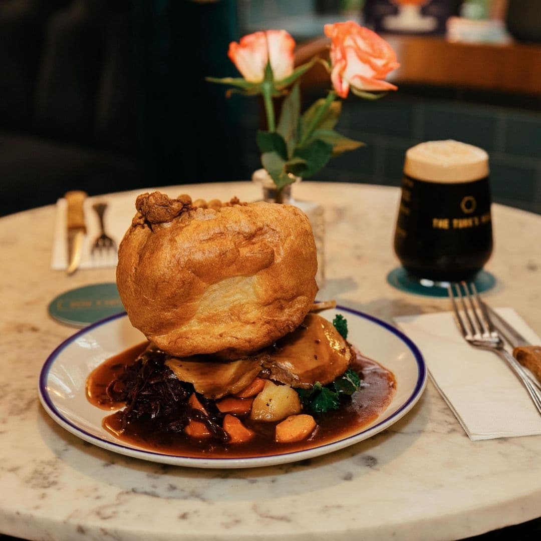 Traditional British Sunday roast with a large Yorkshire pudding, roasted meat, and vegetables, served on a white plate with a pint of dark beer in the background, at The Turk’s Head.