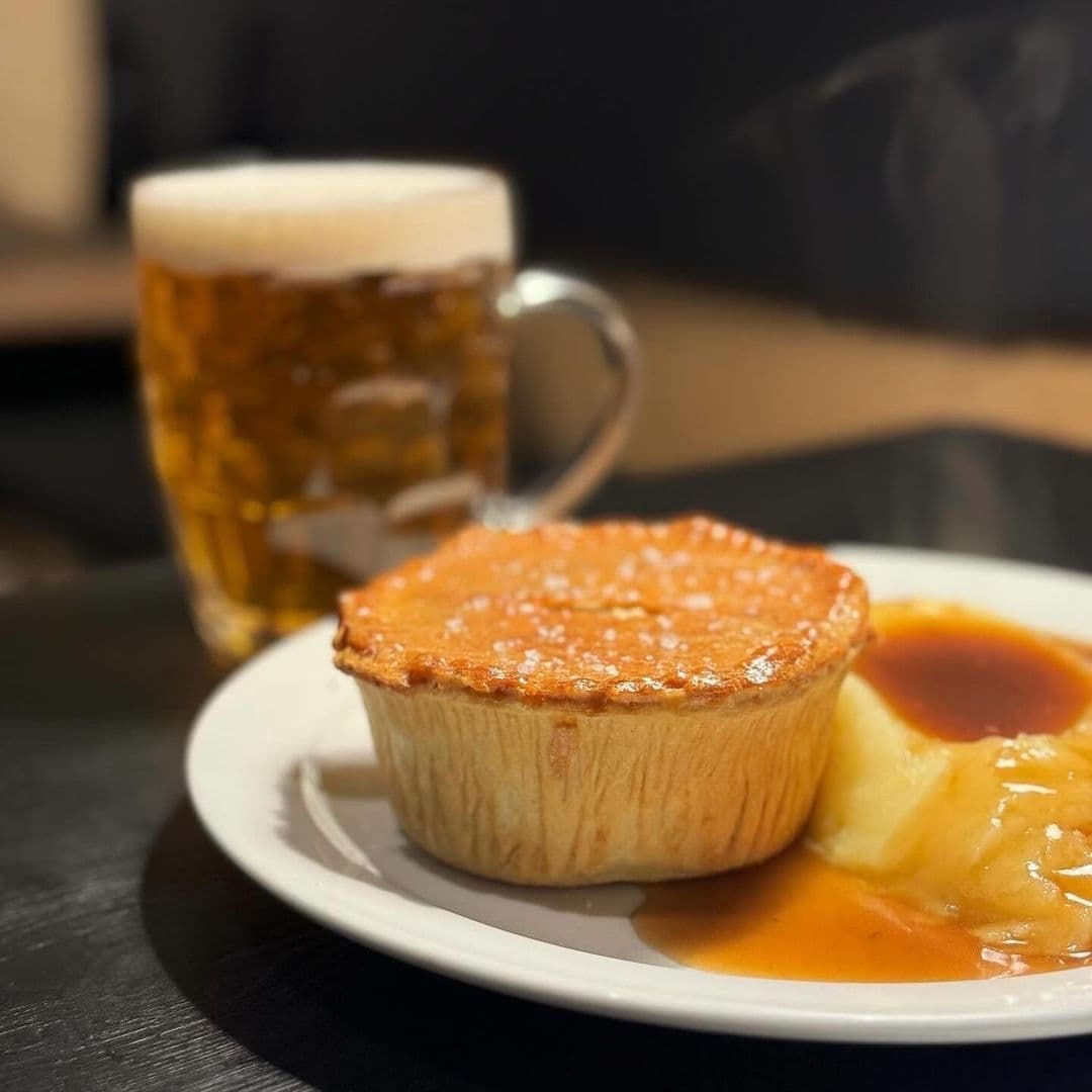 Traditional Australian meat pie with gravy and mashed potatoes on a white plate, accompanied by a pint of beer, at The Melbourne.